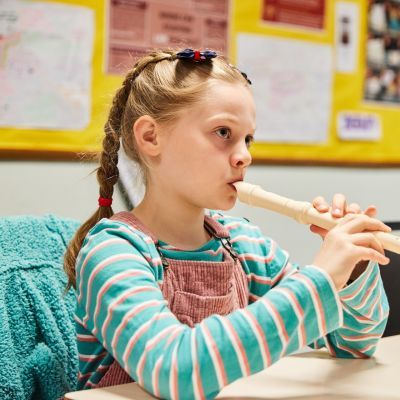 A young girl sits at a desk and plays a flute. She has blonde hair which she wears in two plaits